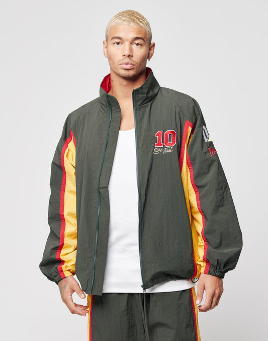 A man with short blond hair wears the LIVE FAST DIE YOUNG Westend Trackjacket—regular fit, multi-logo, in dark green, red, and yellow polyester with 10 on the chest—over a white tank, posing neutrally against a gray background.