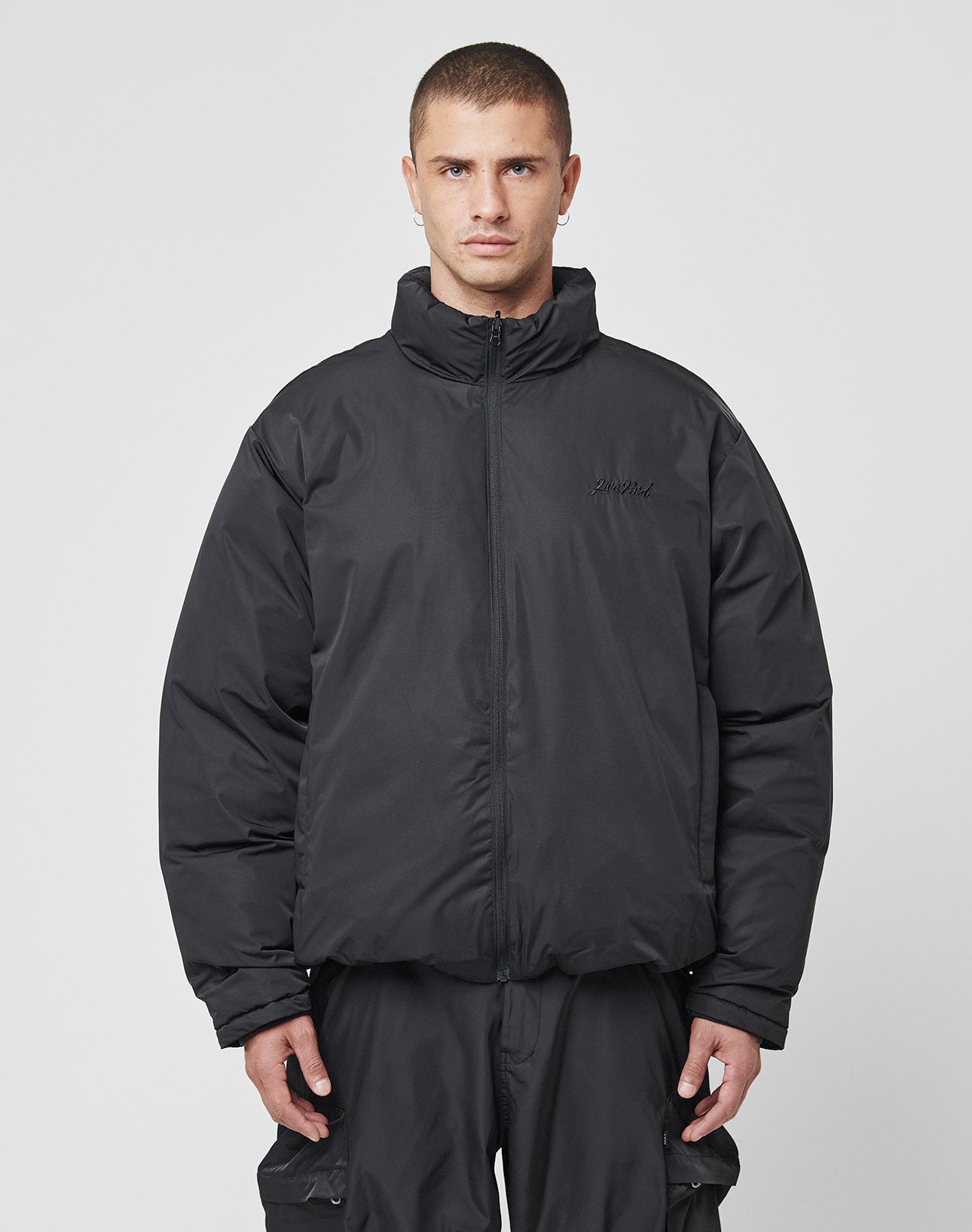 A person faces forward in an oversize fit LFDY Puffer Jacket by LIVE FAST DIE YOUNG with front logo stitching and black cargo pants, set against a plain white background.