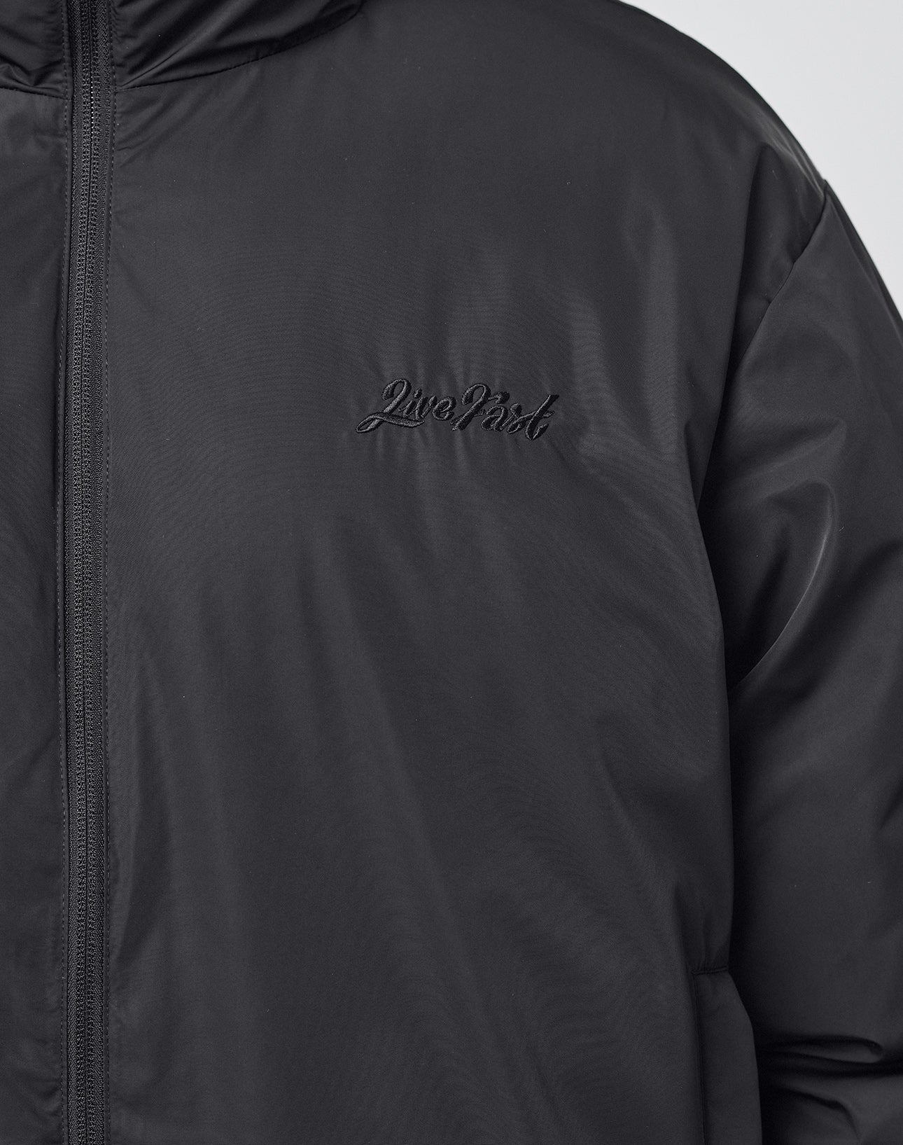 Close-up of the LFDY Puffer Jacket by LIVE FAST DIE YOUNG, featuring black "Live Fast" embroidery on the left chest and smooth, slightly shiny material with a front logo stitch.