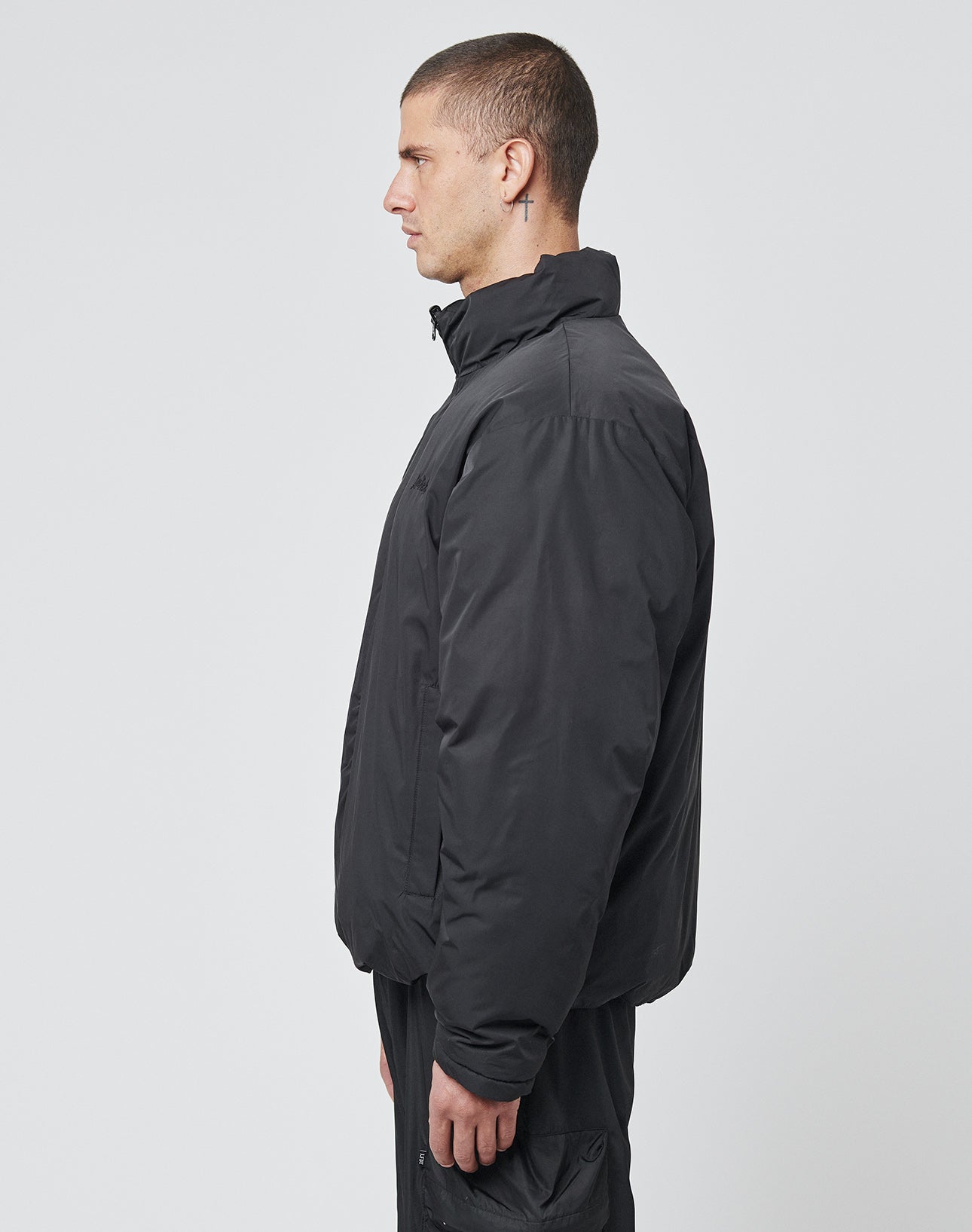 A man with short hair stands in profile facing left against a white background, wearing the LIVE FAST DIE YOUNG LFDY Puffer Jacket in black with front logo stitching and matching black pants.