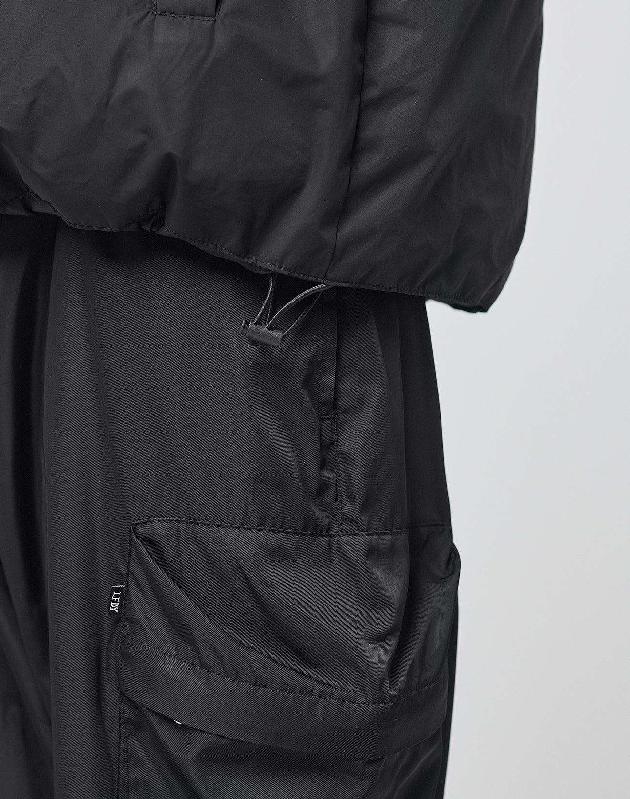 Close-up of a person in a black LIVE FAST DIE YOUNG LFDY Puffer Jacket with front logo stitching and black cargo pants featuring large pockets, highlighting the side and lower jacket details and upper section of the oversize fit pants.