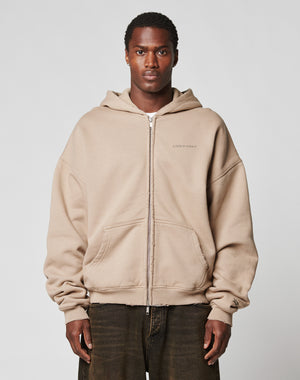 A person stands facing forward in a beige LIVE FAST DIE YOUNG LFDY Double Hooded Zip with subtle waffle lining and front pockets, paired with dark brown pants against a plain, light background.
