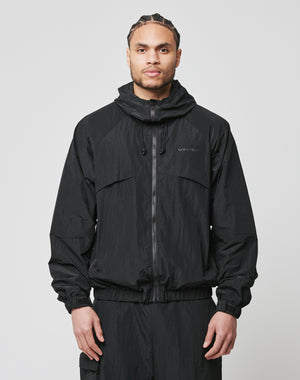 A person stands facing forward, wearing the LIVE FAST DIE YOUNG Live Fast Nylon Jacket in black with seitliche Eingrifftaschen and an oversized fit, paired with matching pants, hands relaxed by their sides against a plain light grey background.