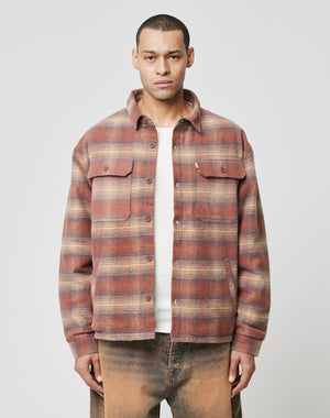 A man faces forward wearing the LIVE FAST DIE YOUNG Vintage Padded Flannel in red and beige plaid over a white shirt and brown pants, set against a plain light gray background.
