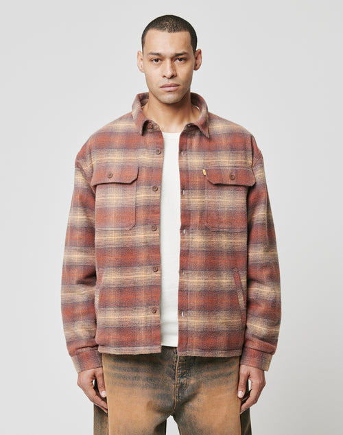 A man faces forward wearing the LIVE FAST DIE YOUNG Vintage Padded Flannel in red and beige plaid over a white shirt and brown pants, set against a plain light gray background.