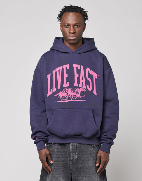 A person wearing the LIVE FAST DIE YOUNG Live Fast Stallion Hooded, an oversize navy cotton hoodie with a pink LIVE FAST horse graphic, stands against a plain light grey background.