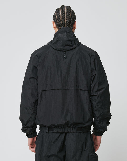 A person with braided hair stands with their back to the camera, wearing a black LIVE FAST DIE YOUNG Live Fast Nylon Jacket with seitliche Eingrifftaschen and oversized black pants against a plain light background.