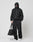 A person in all-black, including a hooded jacket with zipper, face covering, cap, loose pants, and white sneakers, stands against a plain background holding the LIVE FAST DIE YOUNG Live Fast Paintbag.