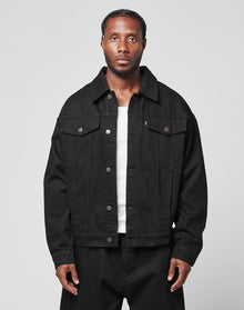 A man with braided hair wears an oversize fit LFDY Denim Jacket by LIVE FAST DIE YOUNG, made from 100% cotton, over a white shirt and black pants. He stands against a plain light gray background, looking directly at the camera with a neutral expression.