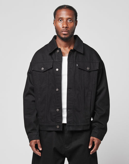 A man with braided hair wears an oversize fit LFDY Denim Jacket by LIVE FAST DIE YOUNG, made from 100% cotton, over a white shirt and black pants. He stands against a plain light gray background, looking directly at the camera with a neutral expression.