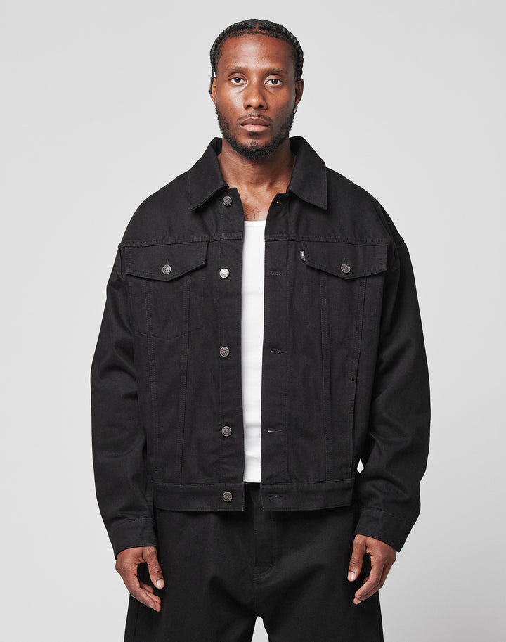 A man with braided hair wears an oversize fit LFDY Denim Jacket by LIVE FAST DIE YOUNG, made from 100% cotton, over a white shirt and black pants. He stands against a plain light gray background, looking directly at the camera with a neutral expression.