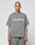 A tattooed, shaved-head individual wears the oversize-fit Basic +49 Tee by LIVE FAST DIE YOUNG, in gray 100% Baumwolle with a bold +49LiveFast front print, against a plain light background.
