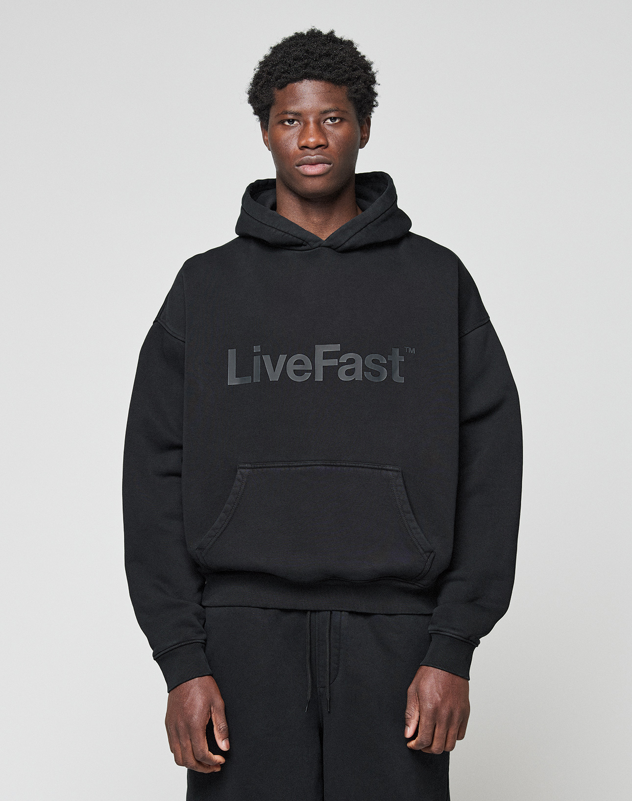 A person wearing the LIVE FAST DIE YOUNG Basic Livefast Hooded in black, featuring a bold "LiveFast" front print and an oversize fit, pairs it with black pants against a plain light gray background.