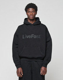 A person wearing the LIVE FAST DIE YOUNG Basic Livefast Hooded in black, featuring a bold "LiveFast" front print and an oversize fit, pairs it with black pants against a plain light gray background.