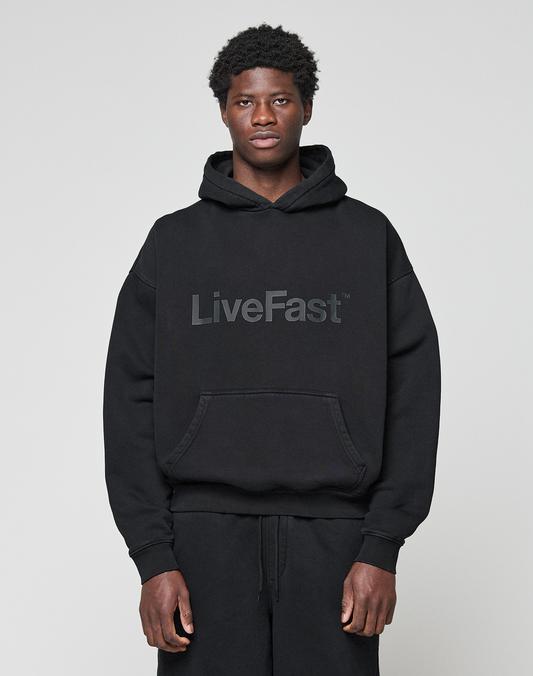 A person wearing the LIVE FAST DIE YOUNG Basic Livefast Hooded in black, featuring a bold "LiveFast" front print and an oversize fit, pairs it with black pants against a plain light gray background.