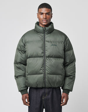A man stands facing forward against a light gray background, wearing the LIVE FAST DIE YOUNG LFDY Puffer Jacket in dark green with an oversize fit and paired with dark jeans.