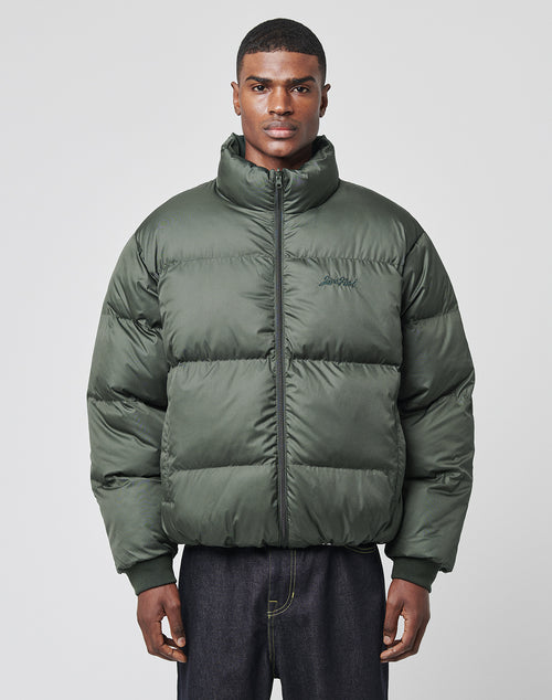 A man stands facing forward against a light gray background, wearing the LIVE FAST DIE YOUNG LFDY Puffer Jacket in dark green with an oversize fit and paired with dark jeans.