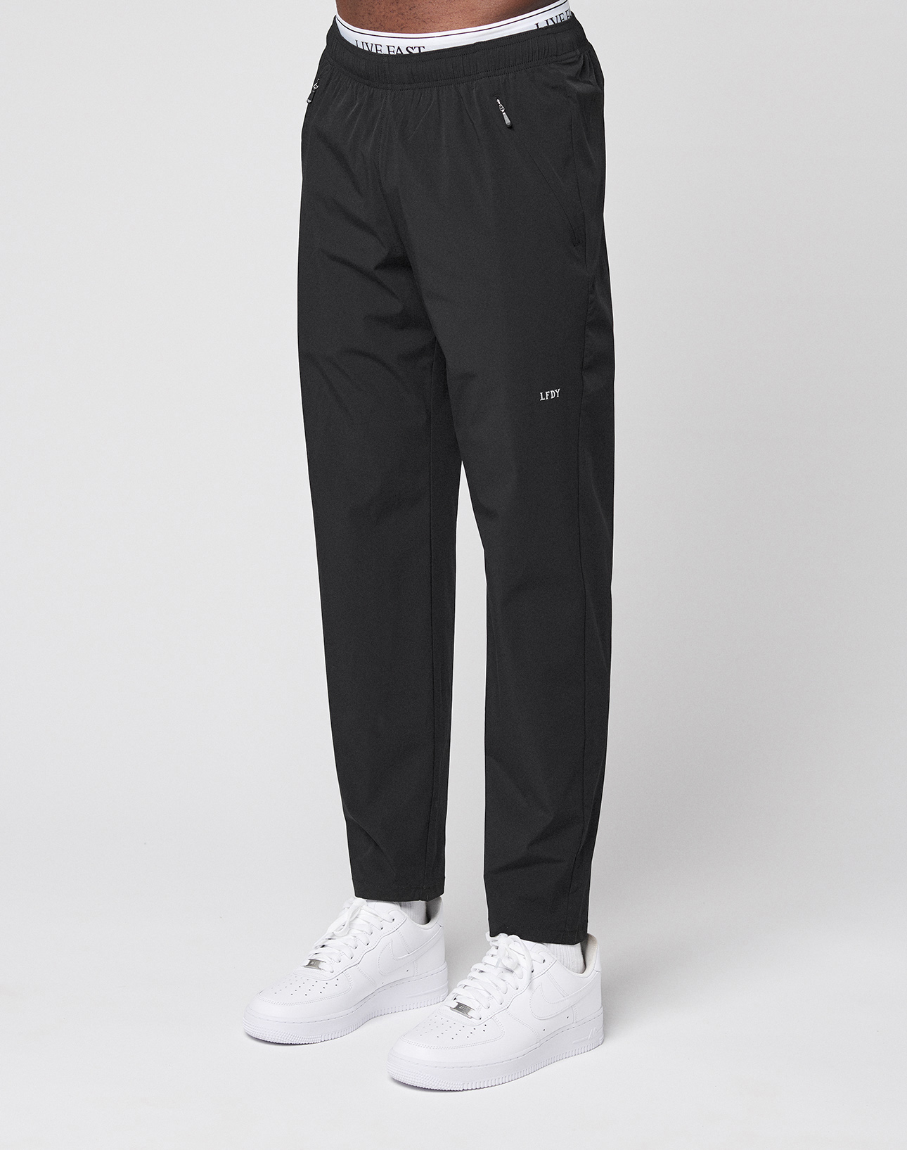 Someone is wearing black LFDY Performance Pants by LIVE FAST DIE YOUNG, featuring an oversize fit, visible branded waistband, and small logo on the thigh, paired with white sneakers and standing against a plain white background.