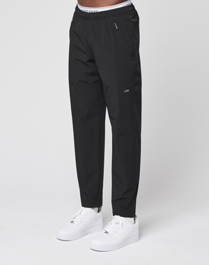 Someone is wearing black LFDY Performance Pants by LIVE FAST DIE YOUNG, featuring an oversize fit, visible branded waistband, and small logo on the thigh, paired with white sneakers and standing against a plain white background.