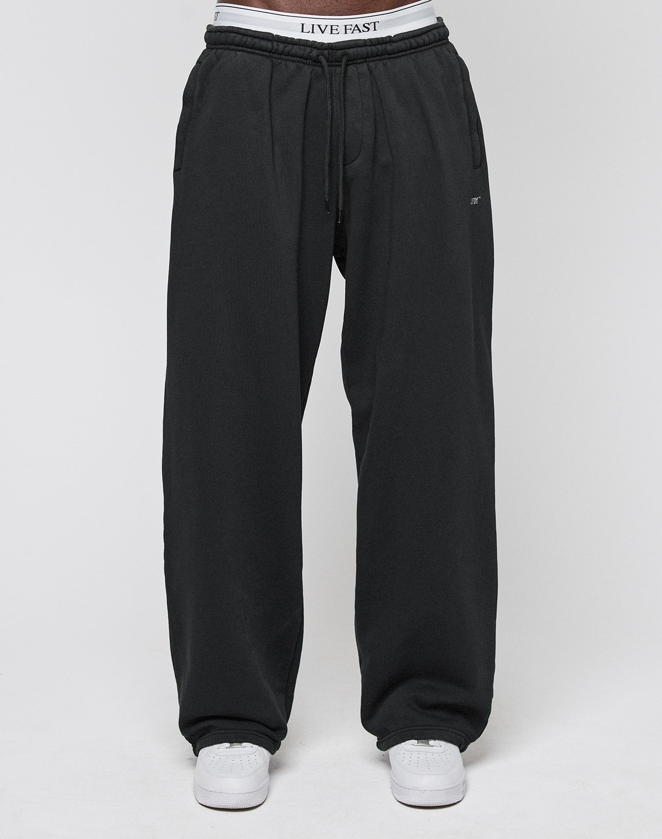 A person wearing the Basic Wide Jogger in black by LIVE FAST DIE YOUNG, featuring an elastic waistband with LIVE FAST text, paired with white sneakers, is standing against a plain white background.