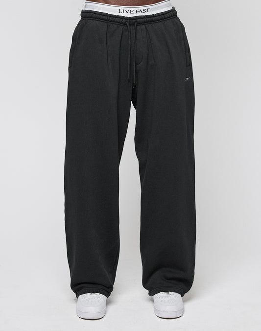 A person wearing the Basic Wide Jogger in black by LIVE FAST DIE YOUNG, featuring an elastic waistband with LIVE FAST text, paired with white sneakers, is standing against a plain white background.