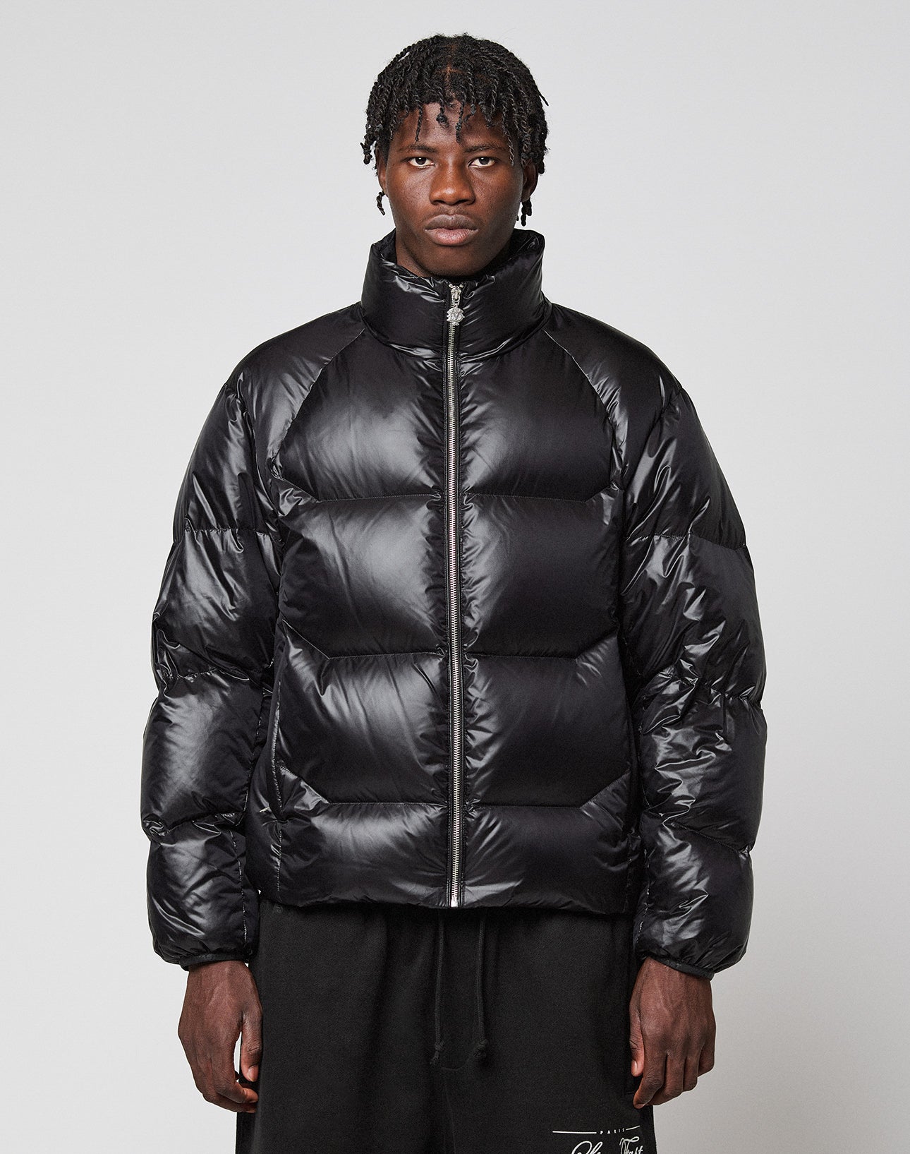 A person with short dreadlocks wears the shiny, wasserabweisend Live Fast Down Jacket by LIVE FAST DIE YOUNG, featuring a high collar, paired with black pants, standing against a plain light gray background.