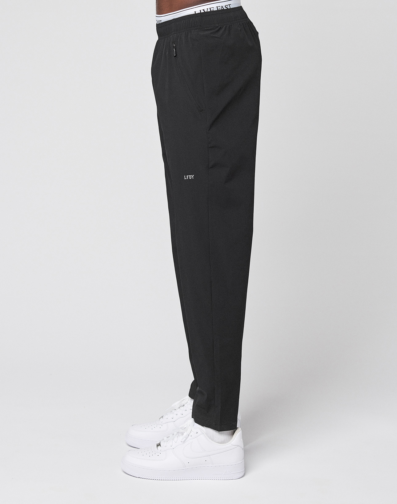 A person stands in profile against a plain light background wearing LIVE FAST DIE YOUNG LFDY Performance Pants in black with an oversize fit and LFYT branding, paired with white sneakers and a white waistband featuring LFYT TASTE text.