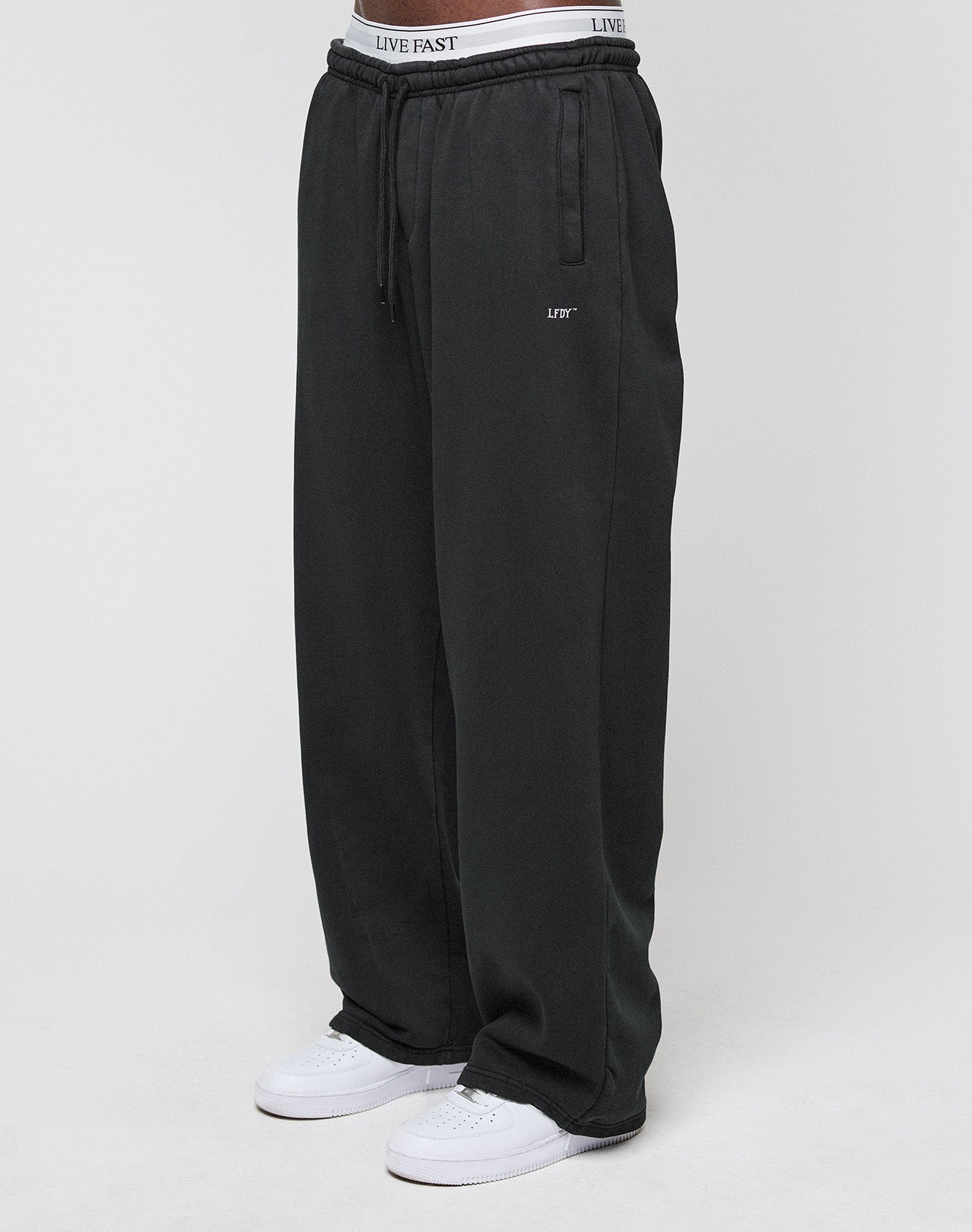 A person wears the LIVE FAST DIE YOUNG Basic Wide Jogger in black with a drawstring waist, white sneakers, and visible LIVE FAST branded underwear. The background is plain and light-colored.