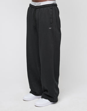 A person wears the LIVE FAST DIE YOUNG Basic Wide Jogger in black with a drawstring waist, white sneakers, and visible LIVE FAST branded underwear. The background is plain and light-colored.