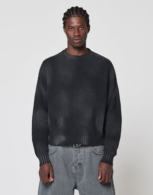 A person with braided hair wears the LIVE FAST DIE YOUNG LFDY Washed Knit Sweater, an oversized dark ribbed piece, paired with baggy gray 100% cotton jeans. They stand against a light gray background, facing forward with a neutral expression.