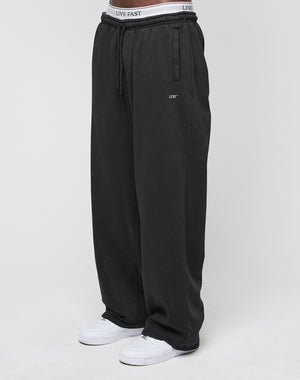 A person wears the LIVE FAST DIE YOUNG Basic Wide Jogger—black, oversize fit with logo print and elastic waistband—paired with white sneakers, standing against a plain white background.