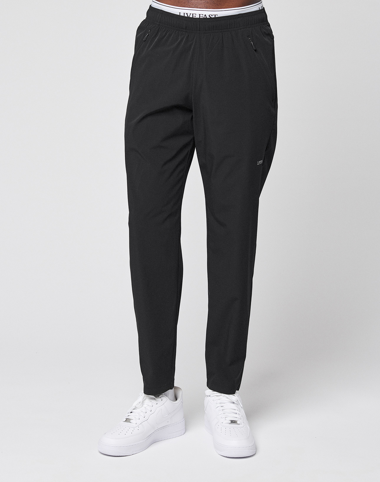 Wearing black LFDY Performance Pants by LIVE FAST DIE YOUNG with zippered pockets, oversize fit, white sneakers, and a LIVE FAST waistband visible on white underwear; subtle LFDY branding stands out against a plain light background.