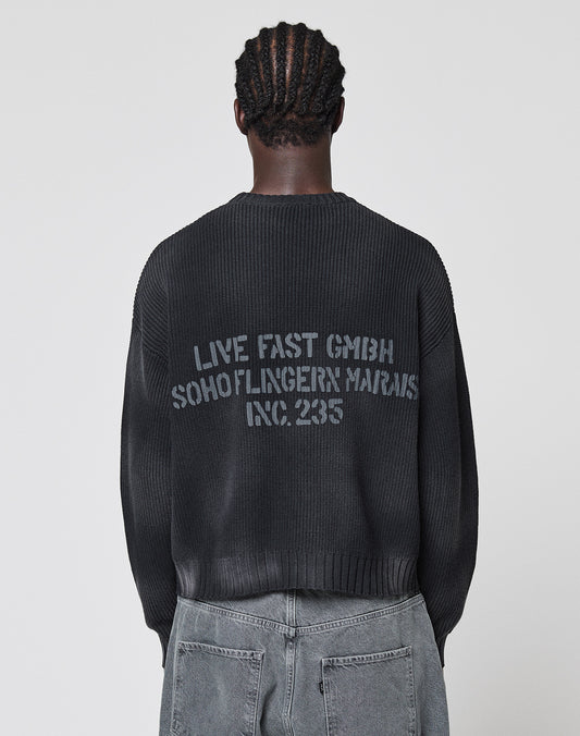 A person with braided hair faces away, wearing the LIVE FAST DIE YOUNG Washed Knit Sweater—an oversized dark ribbed cotton style with a bold back print—paired with faded gray jeans.