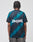 A person with braided hair is seen from behind, wearing the LIVE FAST DIE YOUNG Live Fast Jersey Tee—a black and teal geometric patterned polyester shirt with New York on the back—paired with dark pants.
