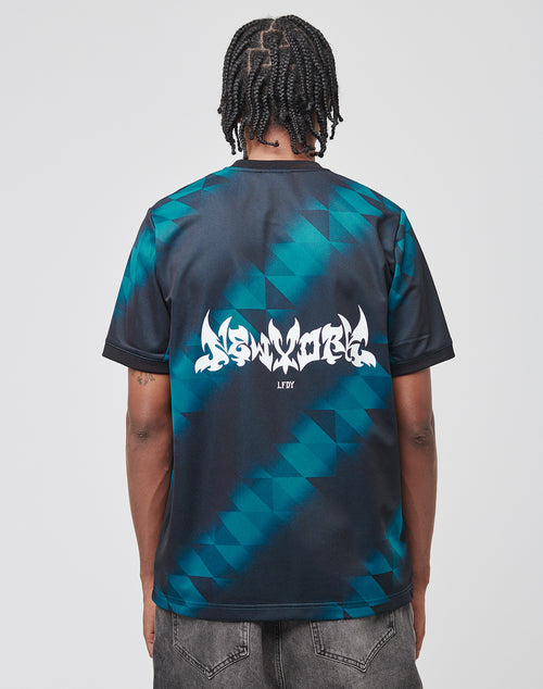 A person with braided hair is seen from behind, wearing the LIVE FAST DIE YOUNG Live Fast Jersey Tee—a black and teal geometric patterned polyester shirt with New York on the back—paired with dark pants.
