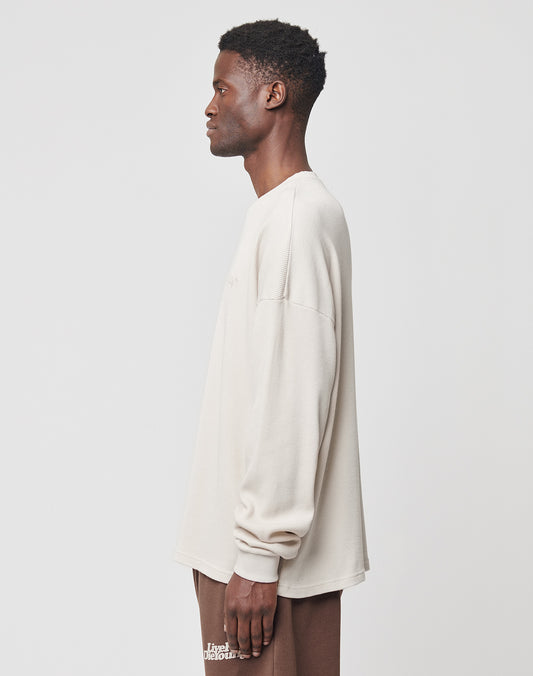 A man stands in profile against a plain background, wearing an oversize beige LIVE FAST DIE YOUNG Basic Waffle Longsleeve and brown pants featuring white Logo-Stick text on the side.