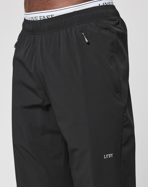 A close-up of a person wearing LFDY Performance Pants by LIVE FAST DIE YOUNG—black, oversized fit, zippered pockets, and “LFDY” logo near the knee. The waistband of white LIVE FAST underwear is visible above the pants.