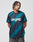 A man with braided hair stands against a plain background, wearing the LIVE FAST DIE YOUNG Live Fast Jersey Tee in black and teal geometric print with white text on the chest and dark jeans. He has a neutral expression and looks slightly downward.