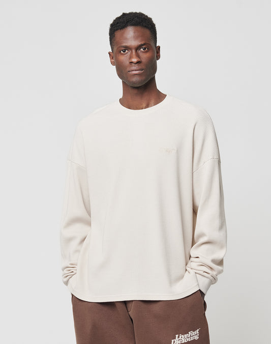 A man wears the LIVE FAST DIE YOUNG Basic Waffle Longsleeve, a cream-colored cotton top in an oversize fit, with brown sweatpants. He stands against a plain white background, hands in pockets, looking at the camera.