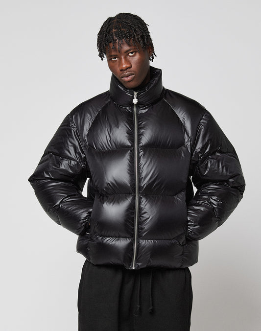 A person with short, twisted hair wears the LIVE FAST DIE YOUNG Live Fast Down Jacket—a shiny, wasserabweisende Oversize-Daunenjacke with 600er Fillpower—standing against a plain light gray background, hands in pockets, and a neutral expression.