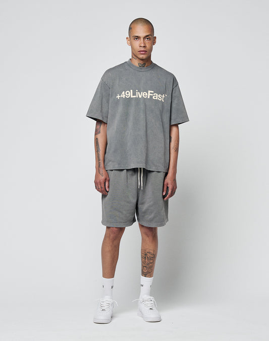 A person wearing the LIVE FAST DIE YOUNG Basic +49 Tee, an oversize 100% cotton grey T-shirt with “+49LiveFast” print, pairs it with grey shorts, white socks, and sneakers. Tattoos are visible on their arms and neck.