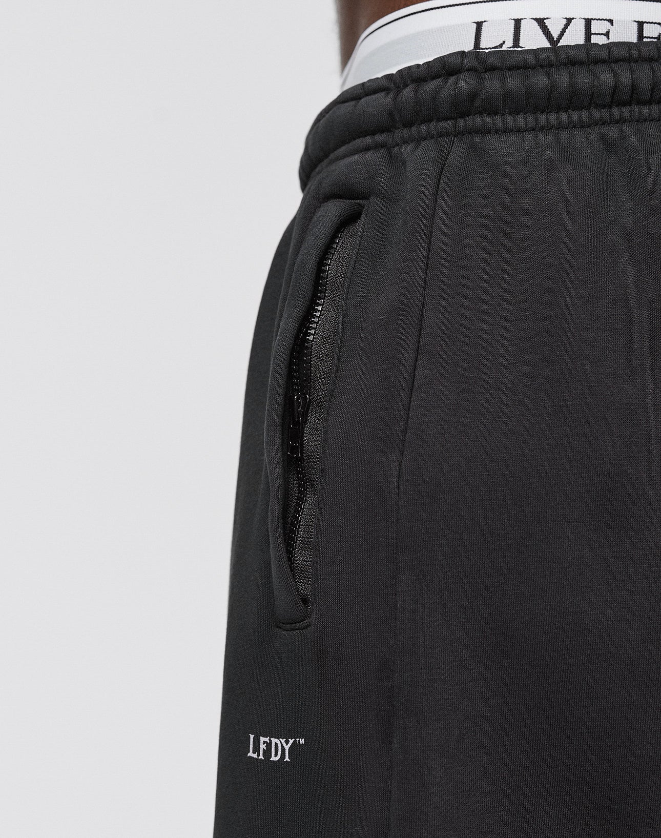 Close-up of a person wearing the Basic Wide Jogger by LIVE FAST DIE YOUNG. The black sweatpants have an elastic waistband with LIVE text, a zippered pocket, and LFDY™ logo print. The background is plain and light-colored.