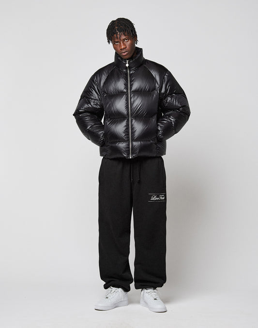 A person stands against a plain background wearing the shiny black Live Fast Down Jacket by LIVE FAST DIE YOUNG with 600er Fillpower, black sweatpants with white script on the upper leg, and white sneakers. Their hands are in their jacket pockets as they look forward.