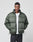 A man stands with his hands in his pockets against a light gray background, wearing the LIVE FAST DIE YOUNG LFDY Puffer Jacket—an oversized green style with front logo stitching—over a white t-shirt.