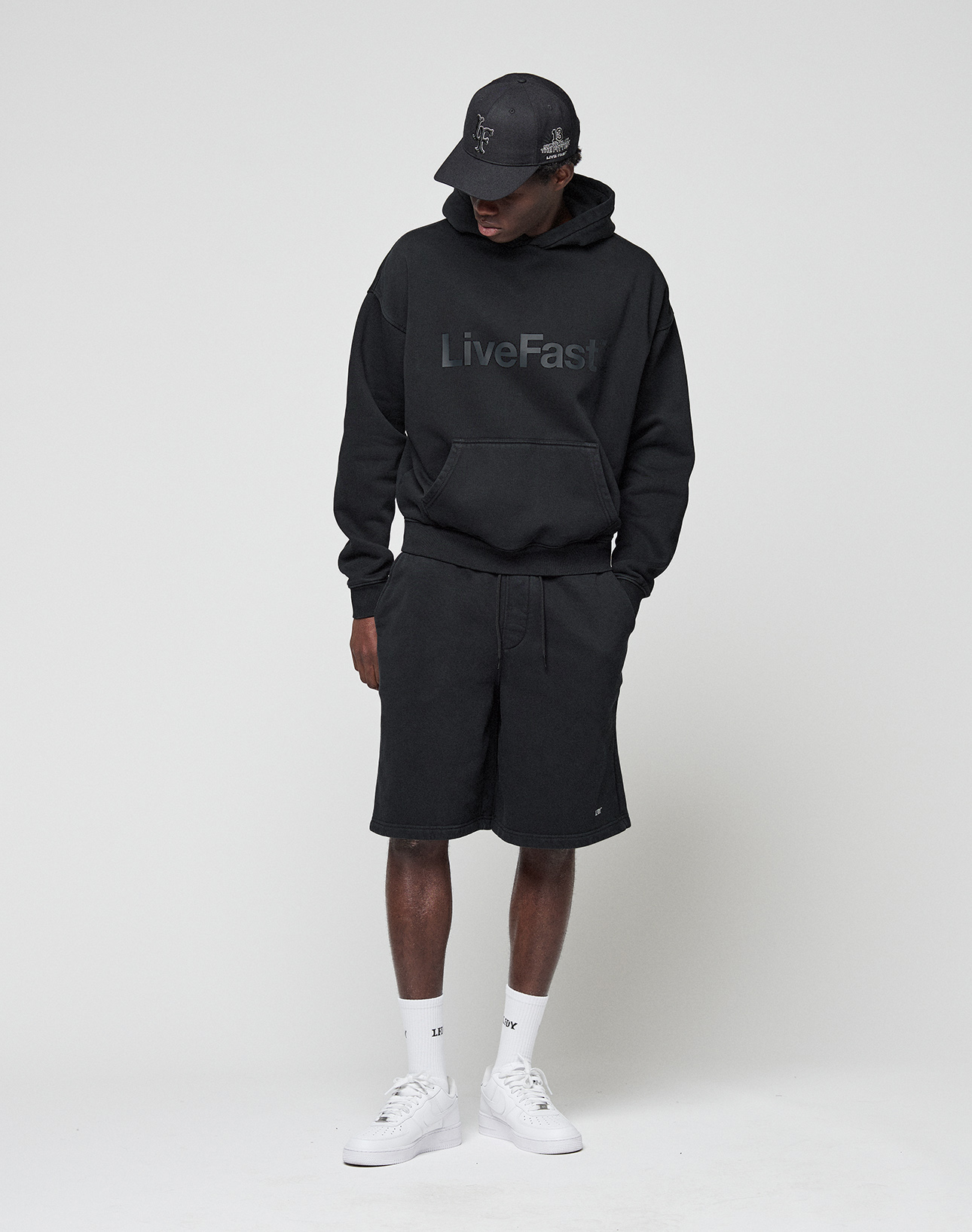 A person in a black LIVE FAST DIE YOUNG LiveFast hoodie with logo print, Basic Shorts with elastic waistband, black cap, white socks, and white sneakers stands against a plain light background, looking down with hands in the hoodie pocket.