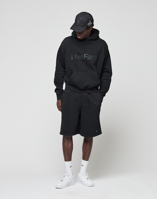 A person in a black LIVE FAST DIE YOUNG LiveFast hoodie with logo print, Basic Shorts with elastic waistband, black cap, white socks, and white sneakers stands against a plain light background, looking down with hands in the hoodie pocket.