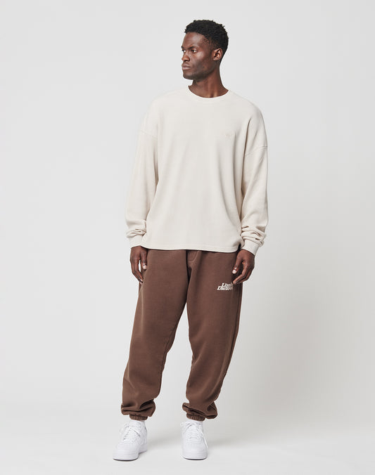 A man stands against a white background wearing the LIVE FAST DIE YOUNG Basic Waffle Longsleeve in cream, brown joggers with a logo patch, and white sneakers. He looks slightly to his left with a neutral expression.