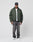 A man in a dark green LIVE FAST DIE YOUNG LFDY Puffer Jacket with front logo stitching, a white tee, loose black jeans, white sneakers, and a black baseball cap stands against a plain light gray background.