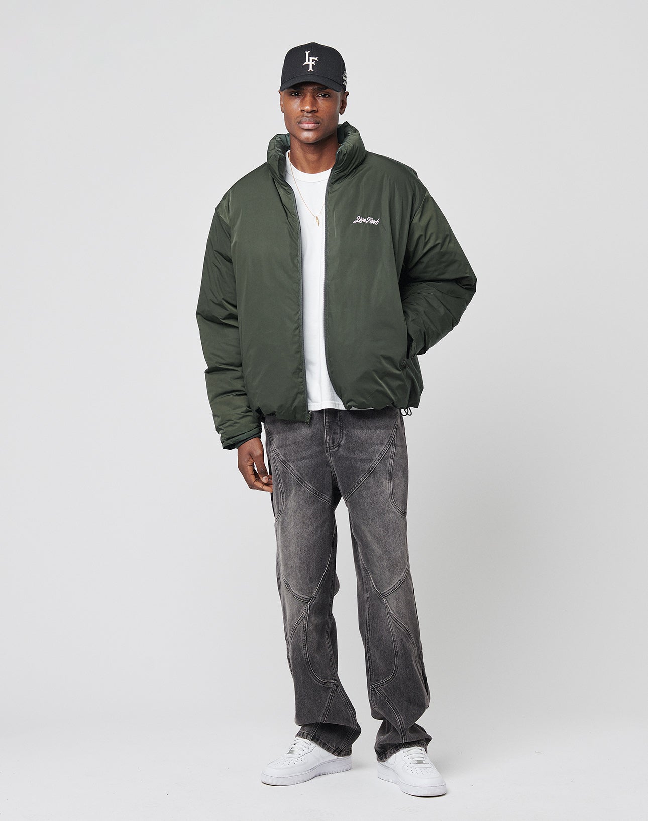 A man stands against a plain light background wearing a black cap, dark green puffer jacket, white t-shirt, LIVE FAST DIE YOUNG Tribal Wide Denim—baggy faded black jeans with white stitching—and white sneakers.