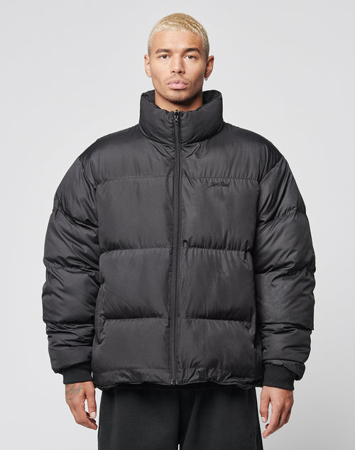 A man with short, bleached blond hair wears the LIVE FAST DIE YOUNG LFDY Puffer Jacket—an oversized black style with front logo stitching and a high collar—while standing against a plain light gray background, gazing directly at the camera.